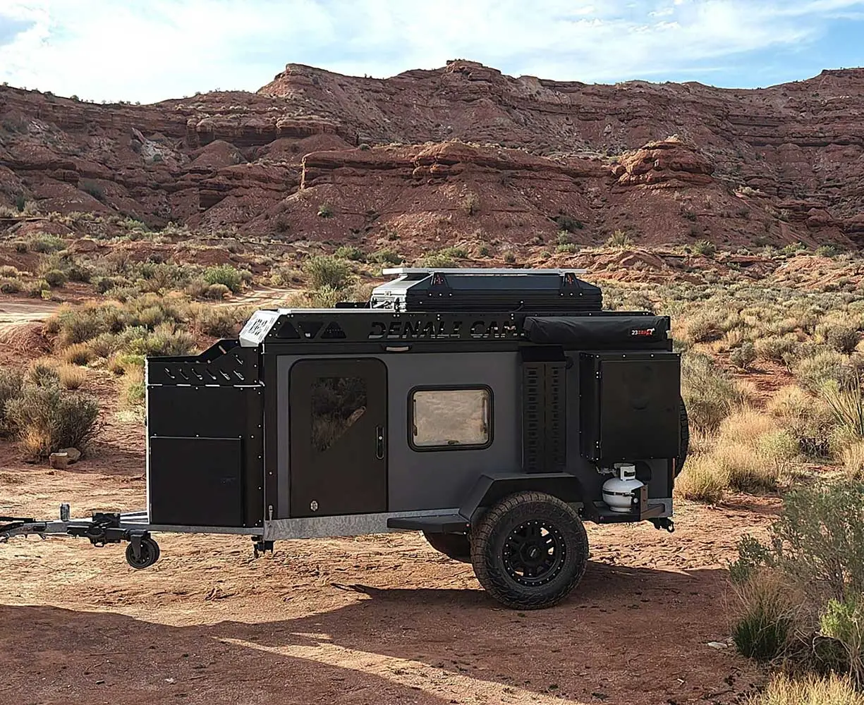 A black off-road camper trailer is parked on a dirt area surrounded by desert plants and red rock formations under a blue sky with scattered clouds.