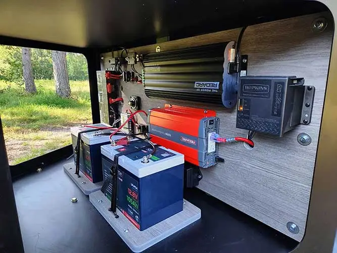 Interior view of a quality electrical setup inside an off-grid camper van featuring two batteries, a red power inverter, a black power converter, and wiring. The background shows a green forest through the open door.