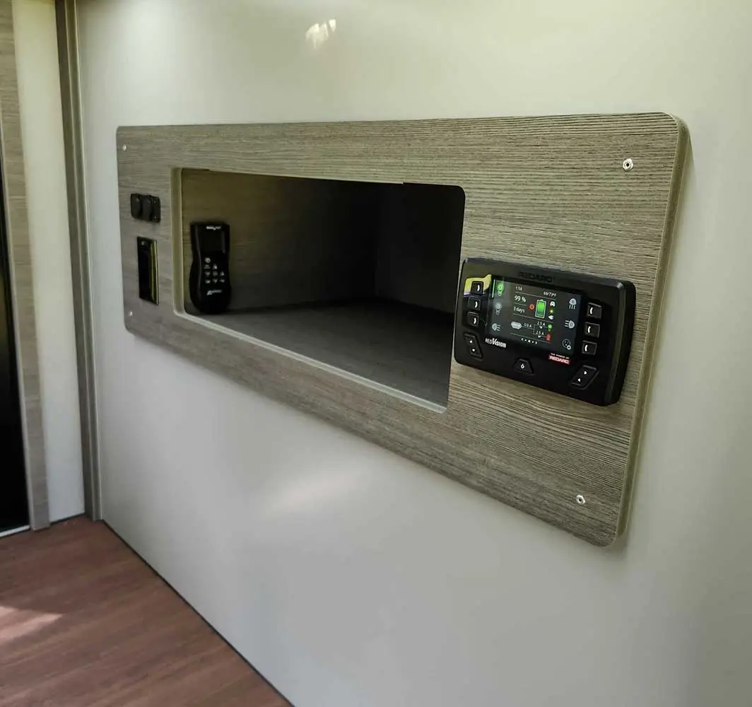 A wall-mounted control panel with a digital display and buttons complements the recessed shelf in a light wood finish, perfect for off-grid living. The shelf holds a small electronic device, all mounted on a light-colored wall above a wooden floor, ideal for modern campers.