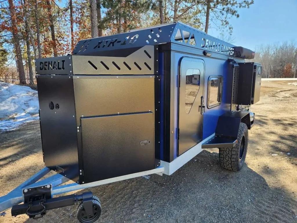 A black and blue Denali camper trailer with rugged tires is parked on a dirt road near trees and patches of snow, featuring storage compartments and a sleek, modern design.