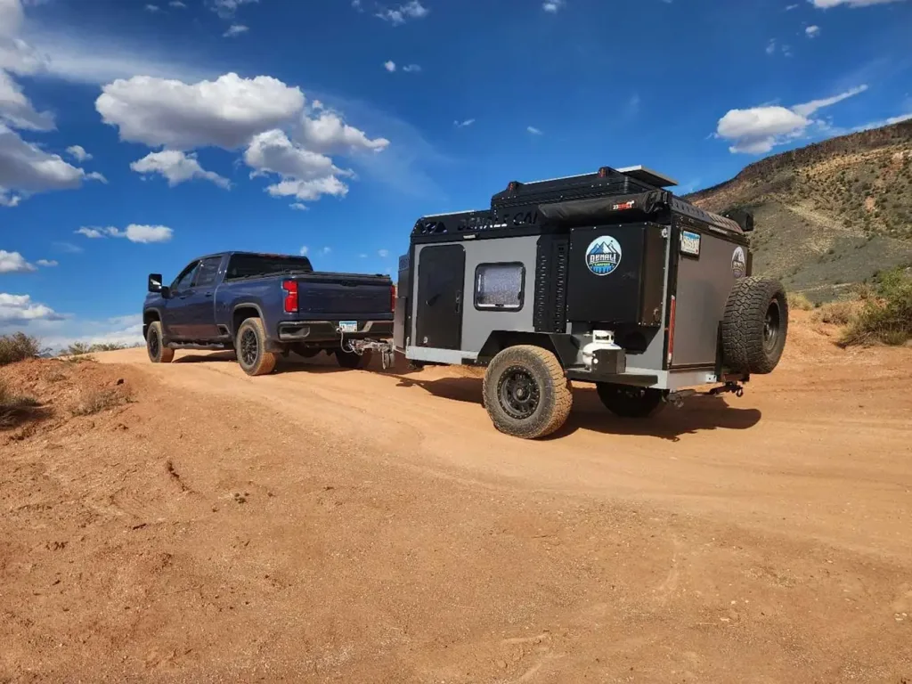 A black pickup truck tows an off-road camper trailer up a dirt hill, under a blue sky with scattered clouds and desert scenery in the background.