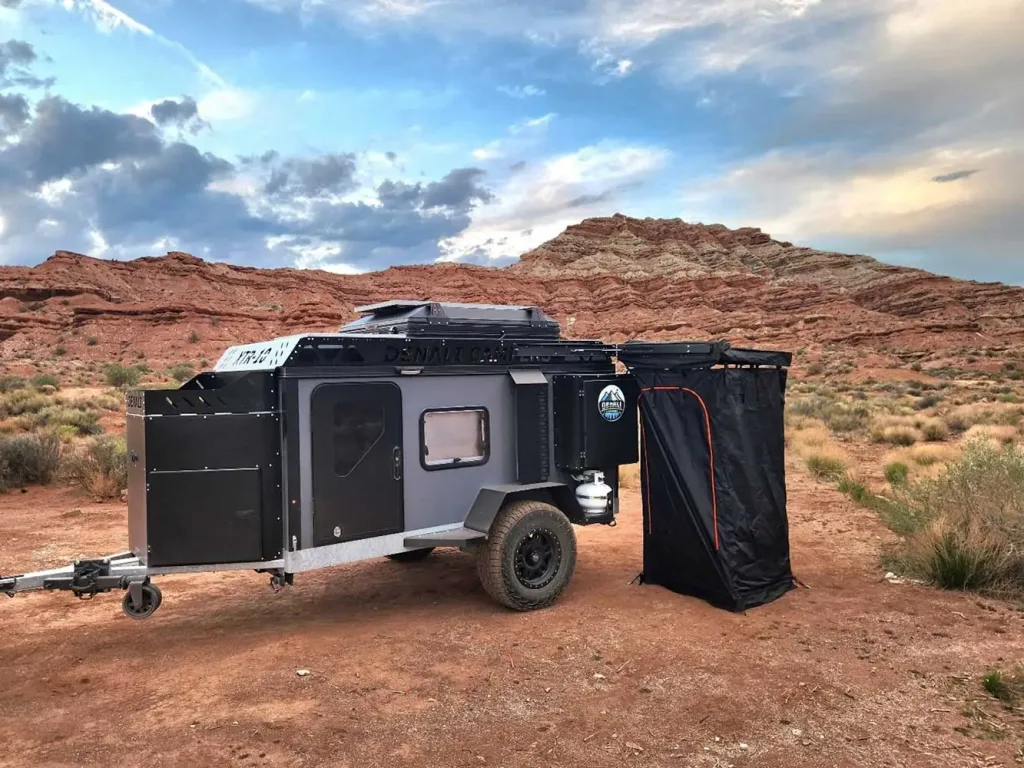 A small off-road camper trailer is parked on desert terrain with red rock formations and cloudy skies in the background. The trailer has an extended awning and an attached outdoor shower area.