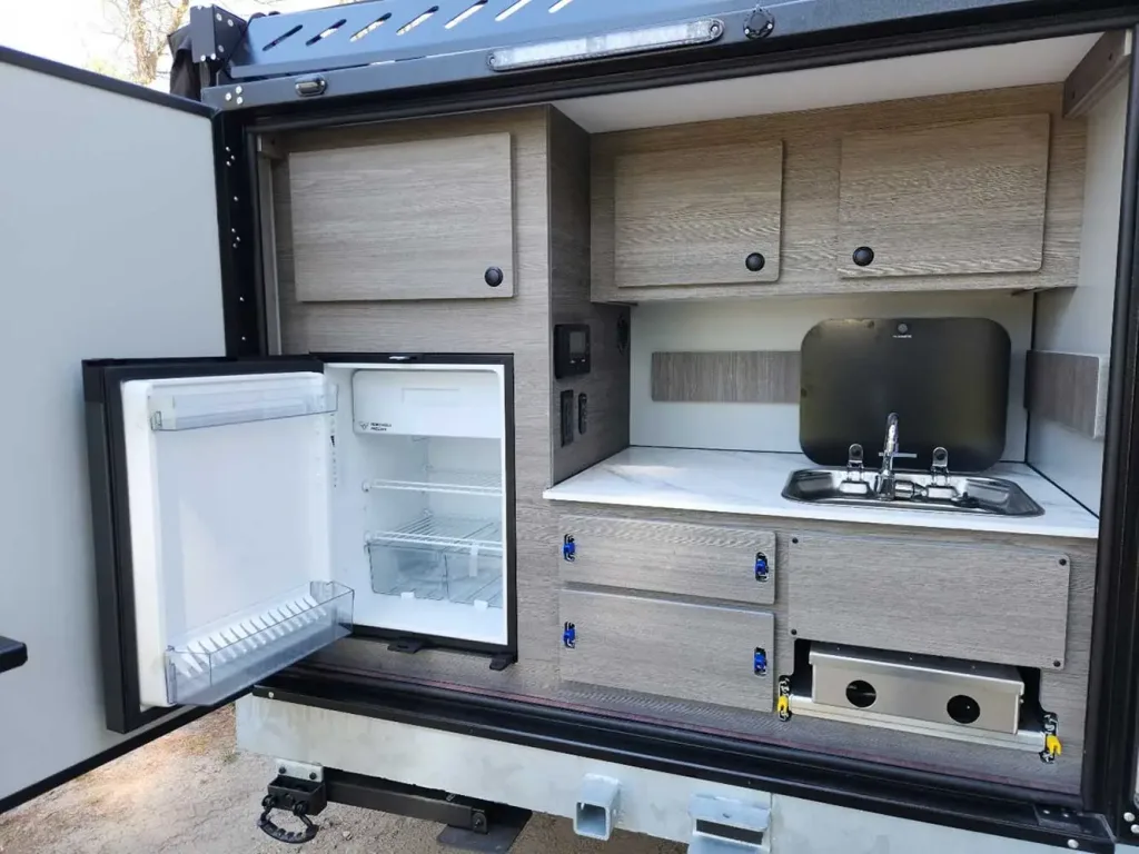A compact outdoor kitchen setup in a vehicle with a small refrigerator, cabinets, a sink with a faucet, a countertop, and a built-in stove below, all in a light wood finish.