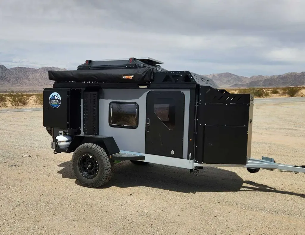 A compact off-road camping trailer with rugged tires and rooftop storage is parked on a desert landscape, with mountains and sparse bushes in the background under a cloudy sky.