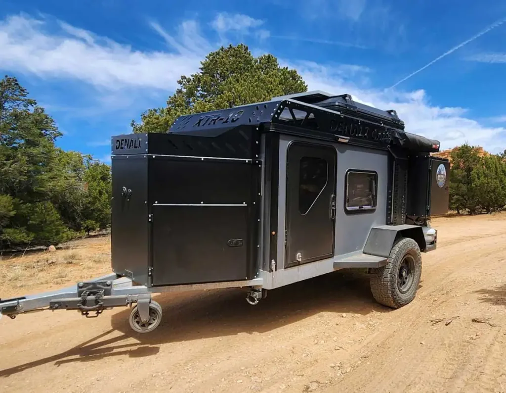 A rugged black and gray off-road camper trailer with large tires is parked on a dirt road surrounded by trees under a blue sky with wispy clouds—perfect inspiration if you're searching for a buying guide before your own adventure.