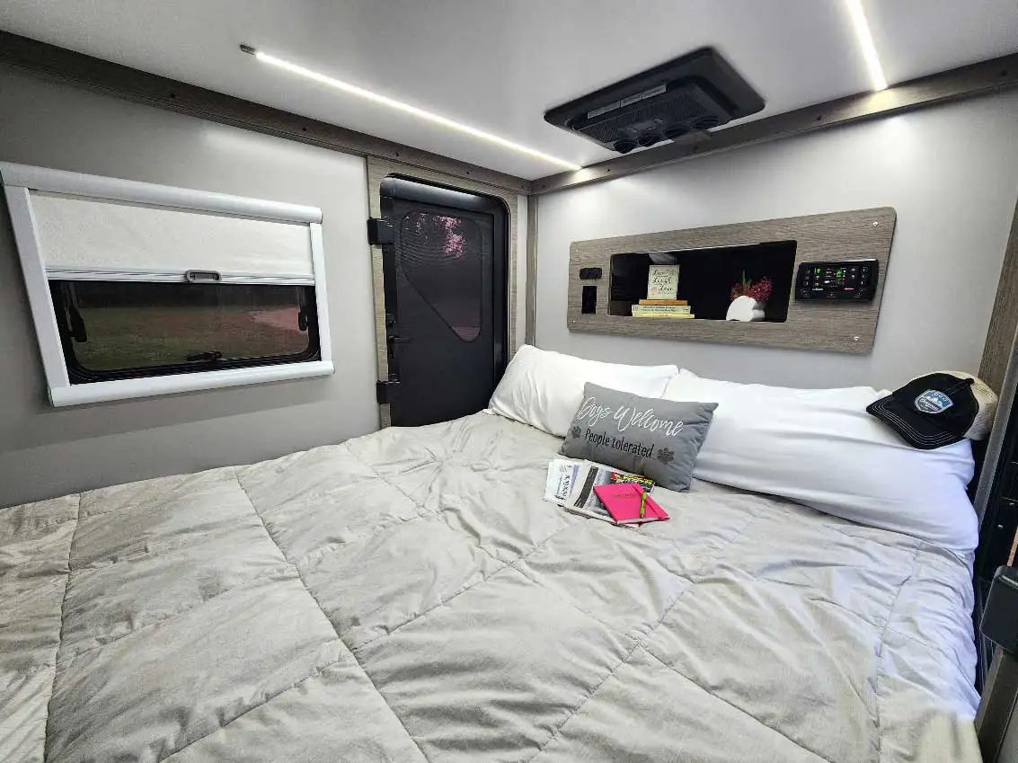 A neatly made bed in a modern RV with a gray comforter, pillows, a hat, and books. There’s a window with a shade, a door, and a shelf above the bed with books and decorations. The decor is cozy and inviting.