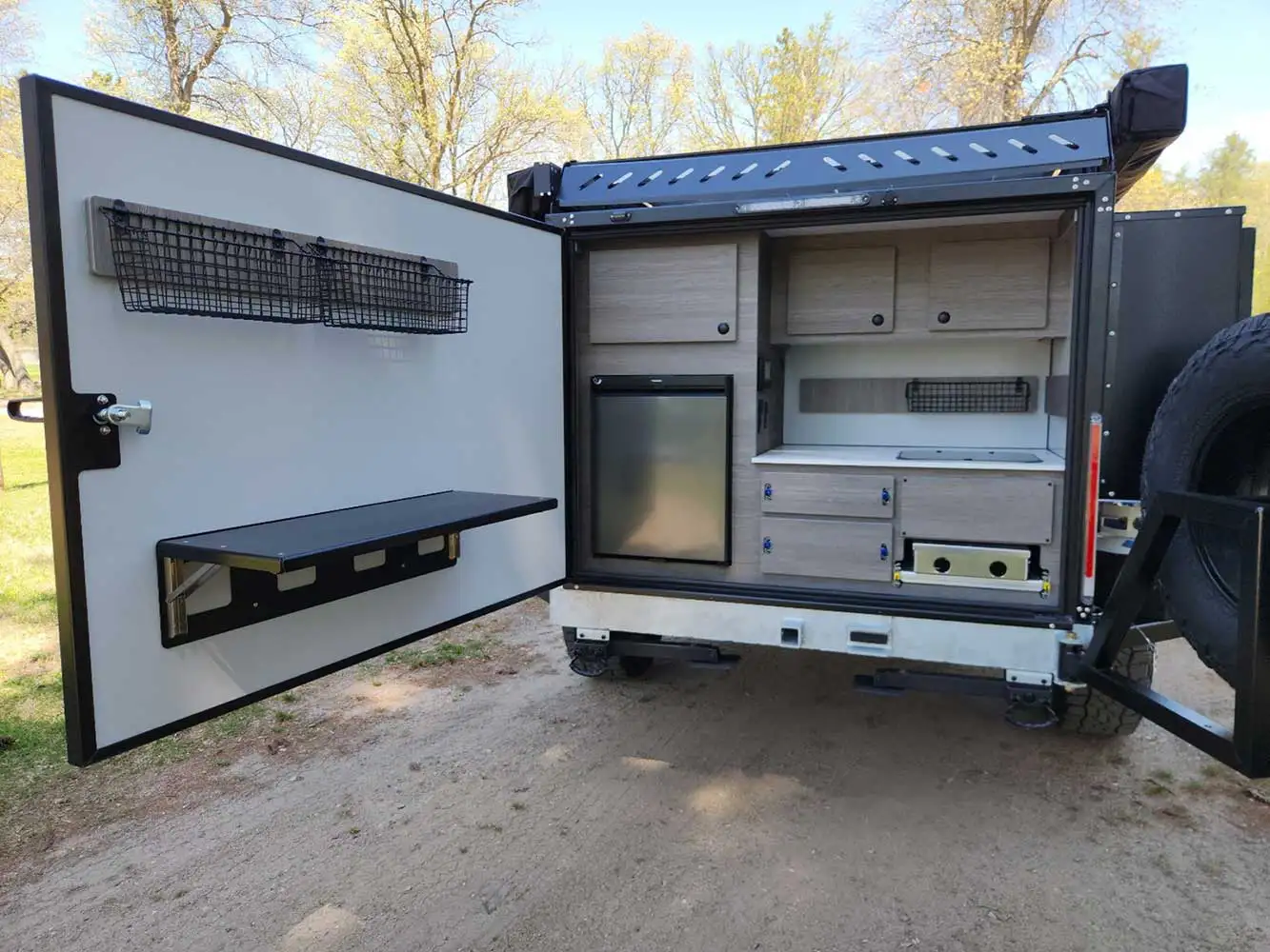 A compact outdoor camper kitchen setup with a fold-down table, small fridge, cabinets, drawers, and shelves, built into the back of an off-road vehicle. The rear door is open, revealing storage and work space.