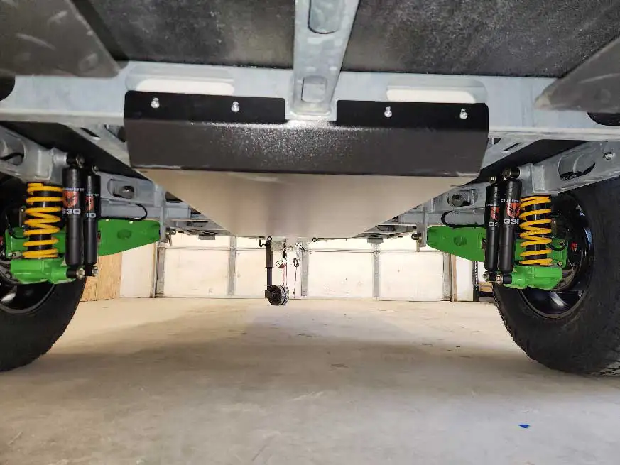 View from underneath a vehicle showing a clean undercarriage, green suspension components with yellow springs, large black tires, and a smooth protective panel, all inside a garage.