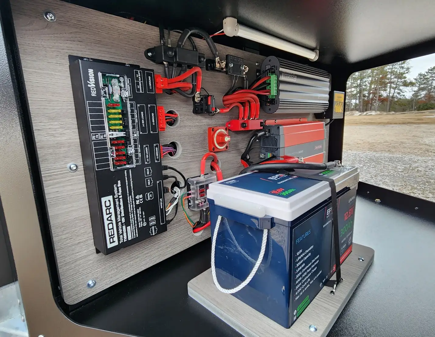 A neatly arranged off-grid electrical setup inside a compartment, featuring a battery, inverter, power management system, wiring, and fuse blocks, with outdoor scenery visible in the background.