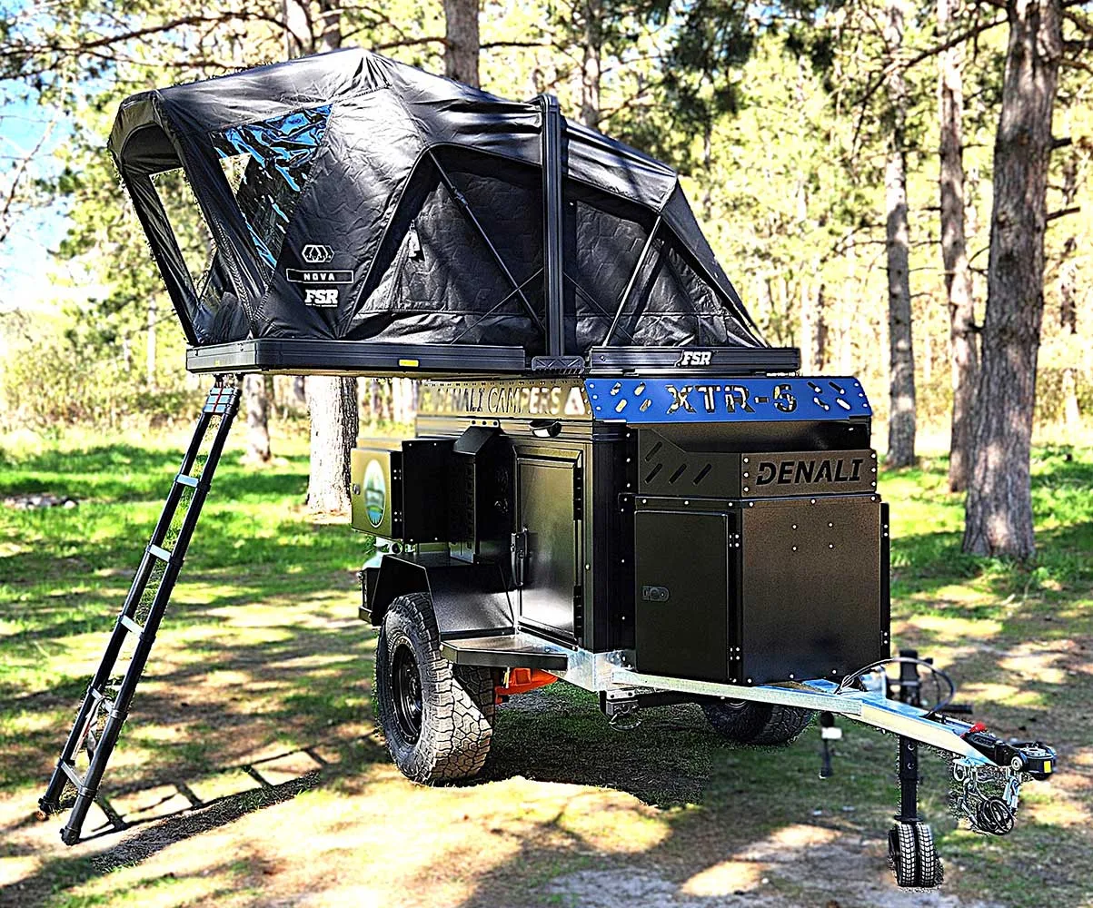 A black off-road camping trailer with raised rooftop tent and ladder is parked on grass in a sunny, wooded area. The trailer has rugged tires and storage compartments labeled “Denali.”.