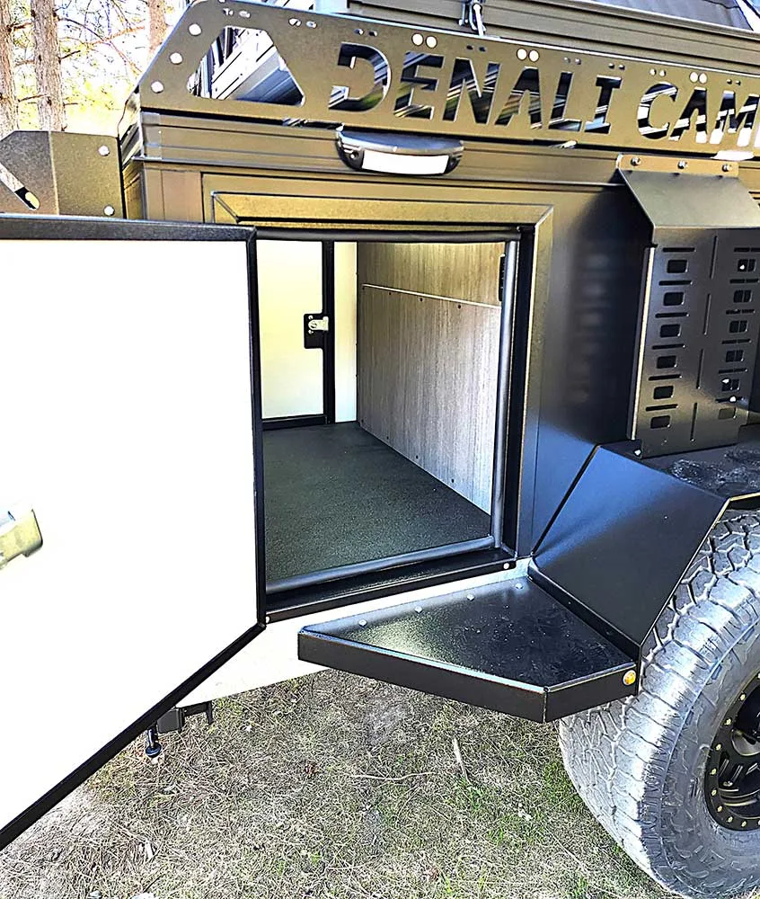 A close-up view of an open camper trailer with a side door revealing a clean, carpeted storage or sleeping area inside; the camper is labeled "Denali Camp" and has rugged, off-road tires.