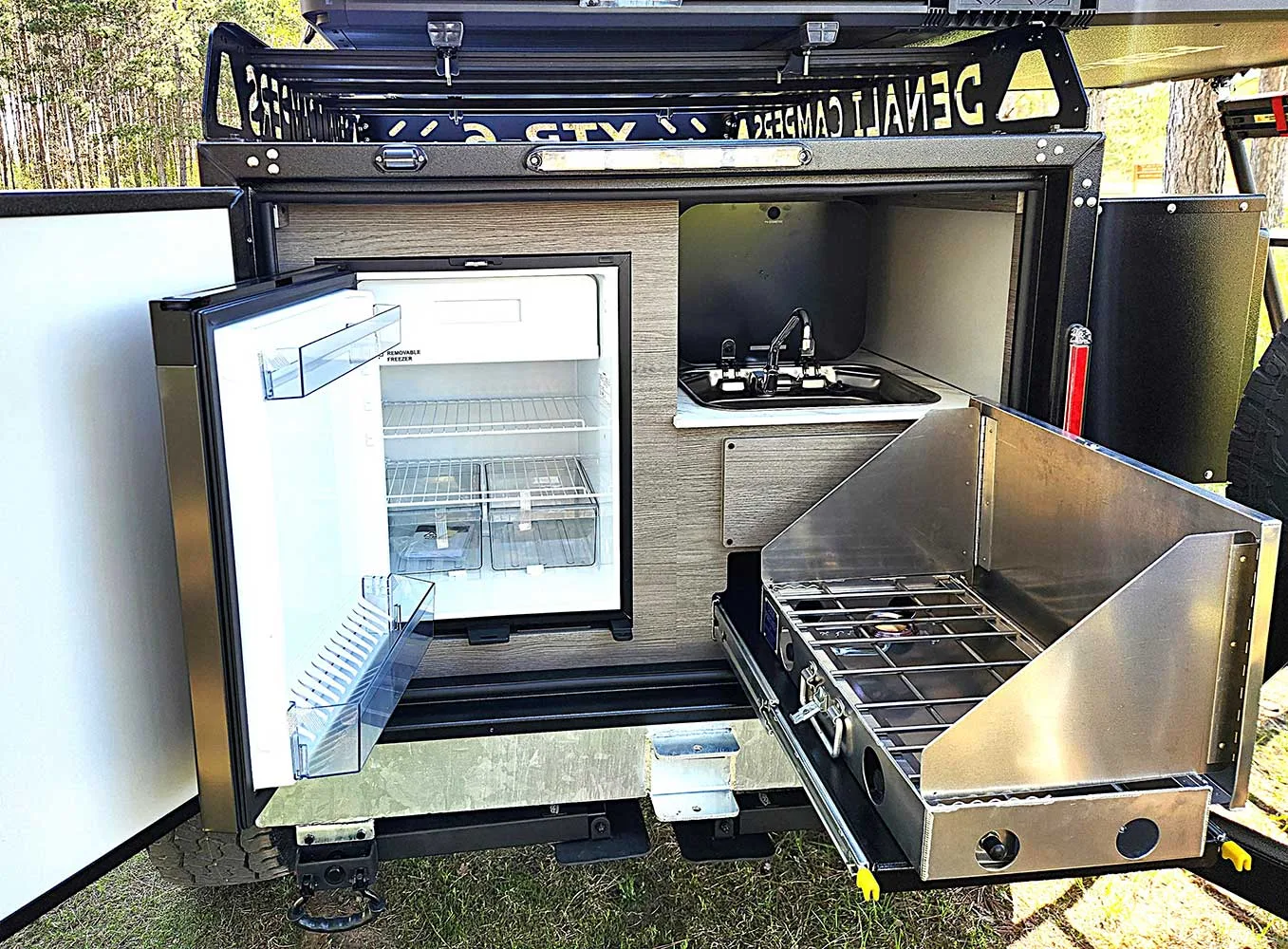 A compact outdoor camping kitchen setup with an open mini fridge, a small sink and stove, and a pull-out dual-burner gas cooktop, all built into a trailer with surrounding greenery visible.
