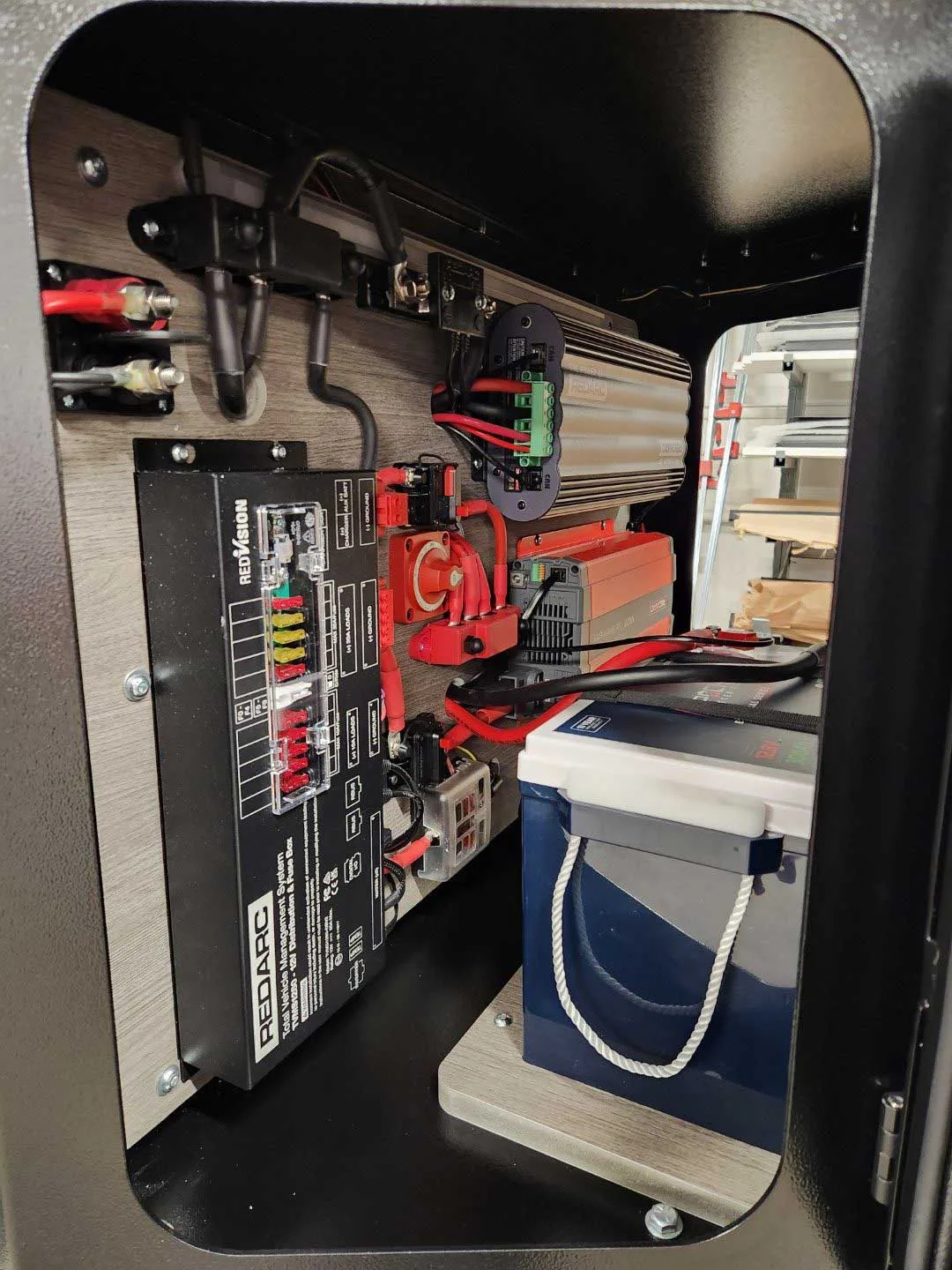 A close-up view of an organized electrical setup featuring a Redarc control box, fuses, wiring, and a large blue battery mounted inside a cabinet with a wood-textured panel.