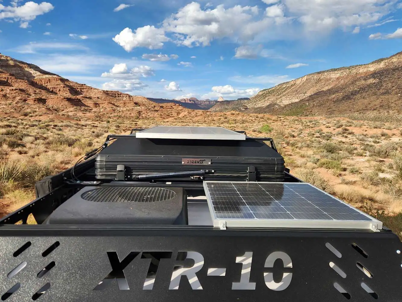 View from the back of an off-road vehicle labeled “XTR-10,” showing solar panels and equipment, parked in a desert landscape with mountains and a partly cloudy blue sky in the background.