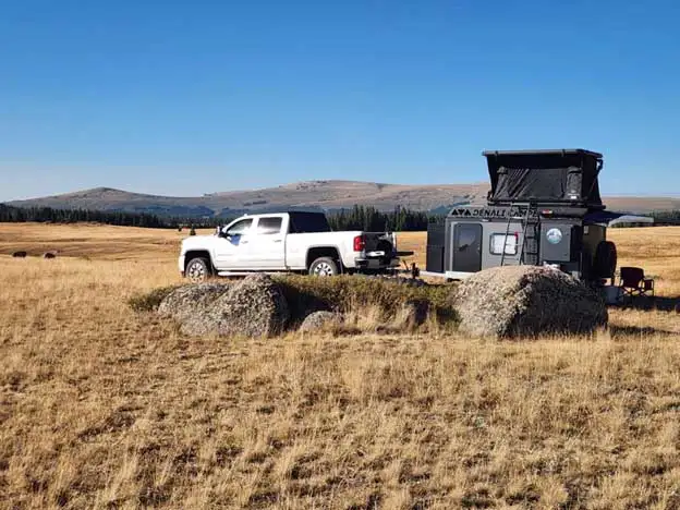 A white pickup truck is parked next to a black camper trailer in an open, grassy field with rocks nearby. Hills and trees are visible in the background under a clear blue sky.