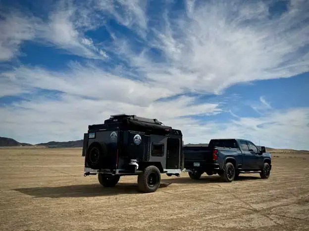 A dark pickup truck towing a rugged off-road camper trailer across a vast, dry desert landscape under a blue sky with scattered clouds.