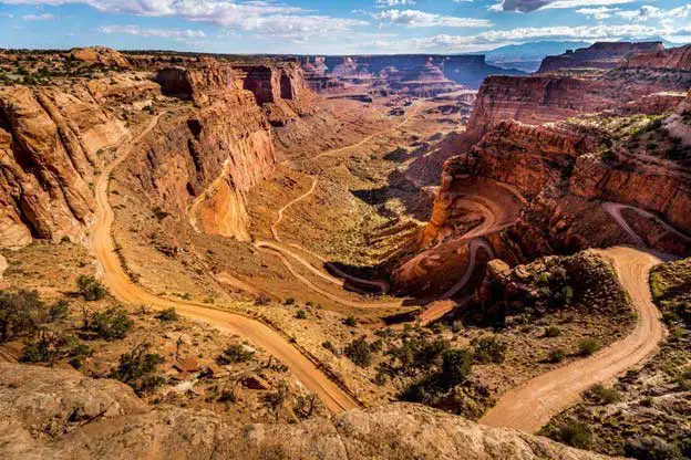 A winding dirt road curves through steep, red rock canyons under a partly cloudy sky, showcasing dramatic cliffs and rugged desert terrain with sparse vegetation.