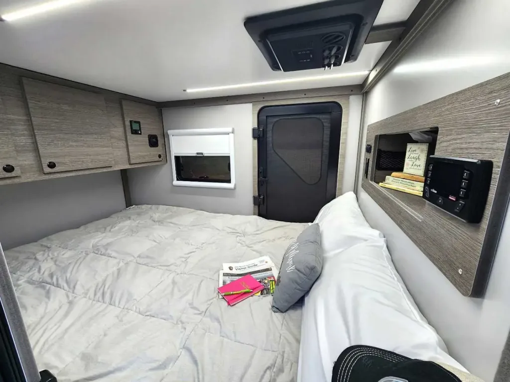 A cozy camper van bedroom with a neatly made bed, white bedding, gray pillow, books on the bed, and overhead cabinets showcases great off grid trailer features like a small window with a blind and a door at the end of the room.