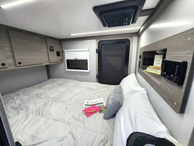 Small camper interior with a neatly made bed, gray bedding, overhead cabinets, and a window with a closed blind—perfect for those exploring the pros and cons of a squaredrop trailer or learning how to choose their ideal mobile space.