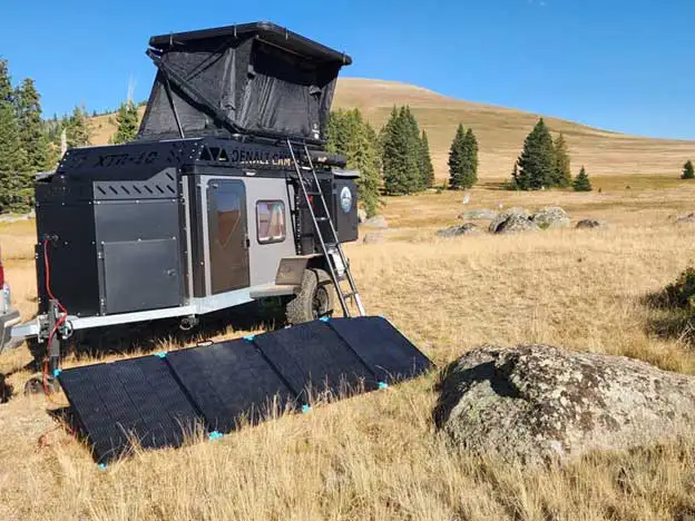 A rugged Squaredrop Trailer with a rooftop tent is parked on dry grass in a remote field. Portable solar panels are set up nearby, while pine trees and rolling hills appear under a clear blue sky.