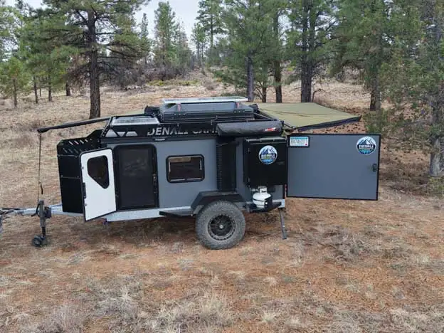 A rugged off-road camping trailer with an extended awning is parked in a forested area of pine trees and dry grass. Outfitted with large tires and Offgrid camper essentials, it’s perfect for true camper life and outdoor adventures.