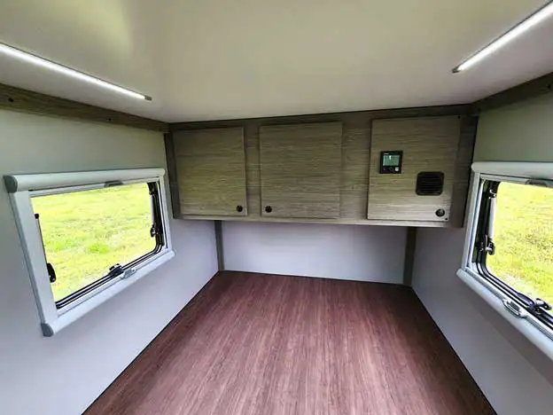 Interior of a small camper with two side windows, wood-like flooring, and upper storage cabinets on the back wall. With views of grassy outdoors, this bright space is perfect for organizing your offgrid camper essentials.