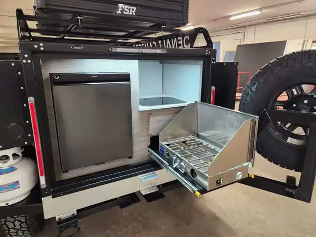 A compact outdoor kitchen setup on a camper trailer, featuring a mini fridge, sink, and pull-out dual-burner stove—true offgrid camper essentials next to the mounted spare tire and propane tank for life beyond the pavement.