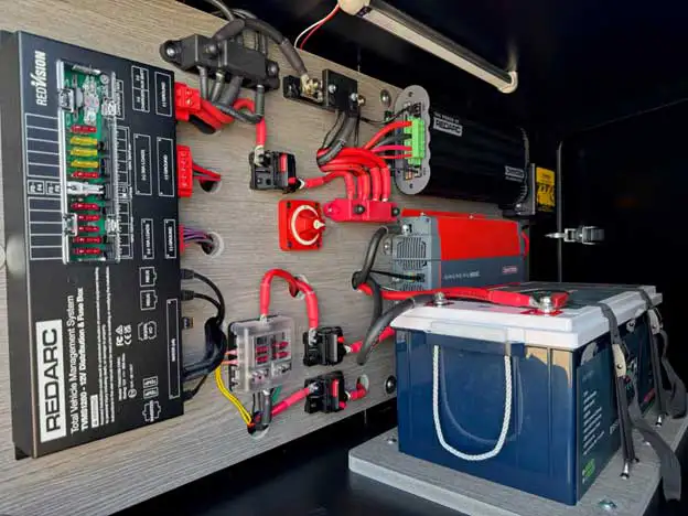 A close-up view of an organized electrical setup featuring a Redarc control panel, various wires, fuses, and a large blue battery—showcasing camper essentials for reliable off grid camping inside a compartment with a wood-textured wall.