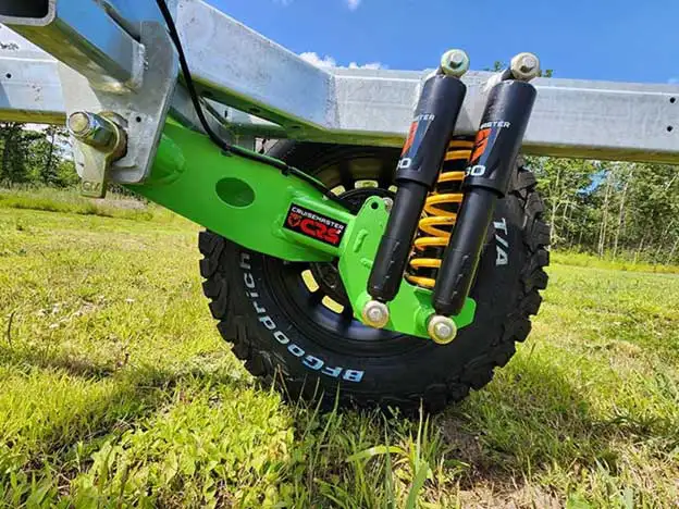 Close-up of a heavy-duty off-road tire with a green suspension arm and dual black shock absorbers, mounted on a metal trailer frame—perfect for life beyond the pavement and offgrid camper essentials—parked on grass with trees and blue sky in the background.