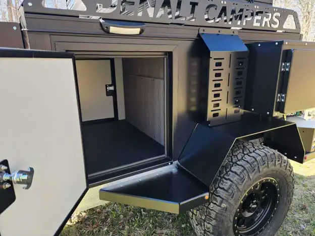 A close-up of a black off-road camper trailer with an open side storage compartment—ideal for storing your offgrid camper essentials—rugged tire, and "Denali Campers" sign on top. The empty compartment and trailer are parked on grass.