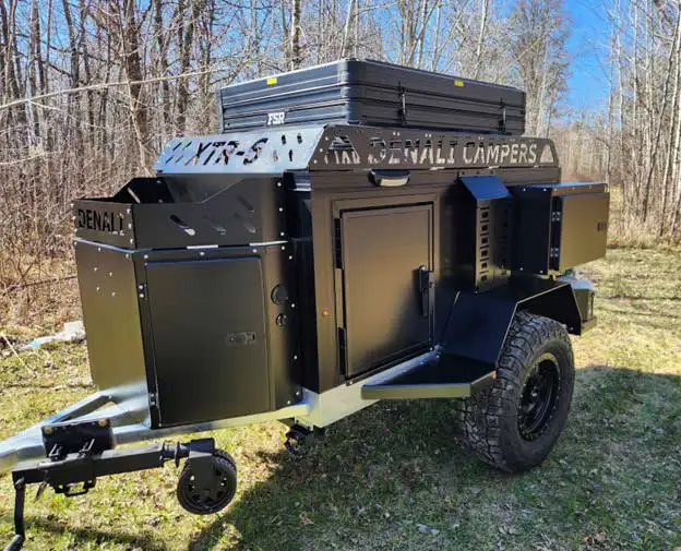 A compact off-road camping trailer with large all-terrain tires, black storage compartments, and a rooftop cargo box—packed with Offgrid Camper Essentials—parked on grassy ground near a forest with leafless trees.