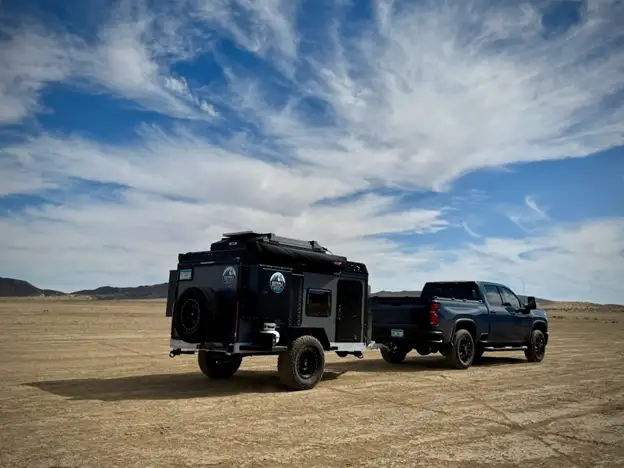 A dark pickup truck tows a purpose-built camper across a dry, flat desert landscape under a blue sky with scattered clouds.