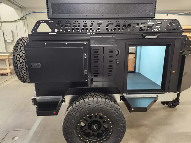 A purpose-built black off road camper trailer with large all-terrain tires, a side-access door leading to a compact interior, and "DENALI CAMPERS" cut into the metal panel above.