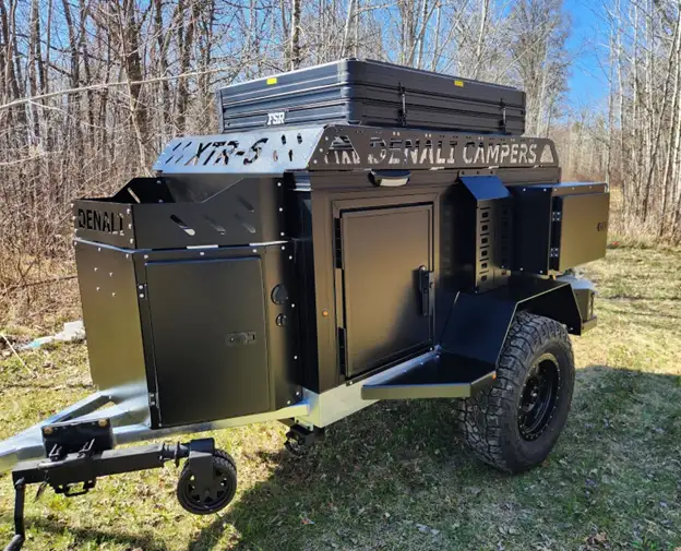 A rugged, black purpose-built Denali off road camper trailer with off-road tires is parked on grass near leafless trees. The boxy camper features storage compartments and equipment mounted on top.