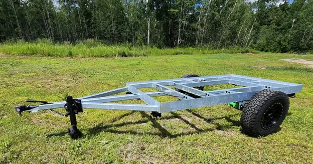 A purpose-built metal utility trailer frame with two black wheels and a tow hitch is parked on grassy ground, surrounded by green trees and vegetation in the background.