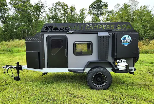 A compact, black and gray Denali Campers purpose-built off road camper is parked on grass, equipped with all-terrain tires, a roof rack, propane tanks, and external storage. Trees and greenery are in the background.