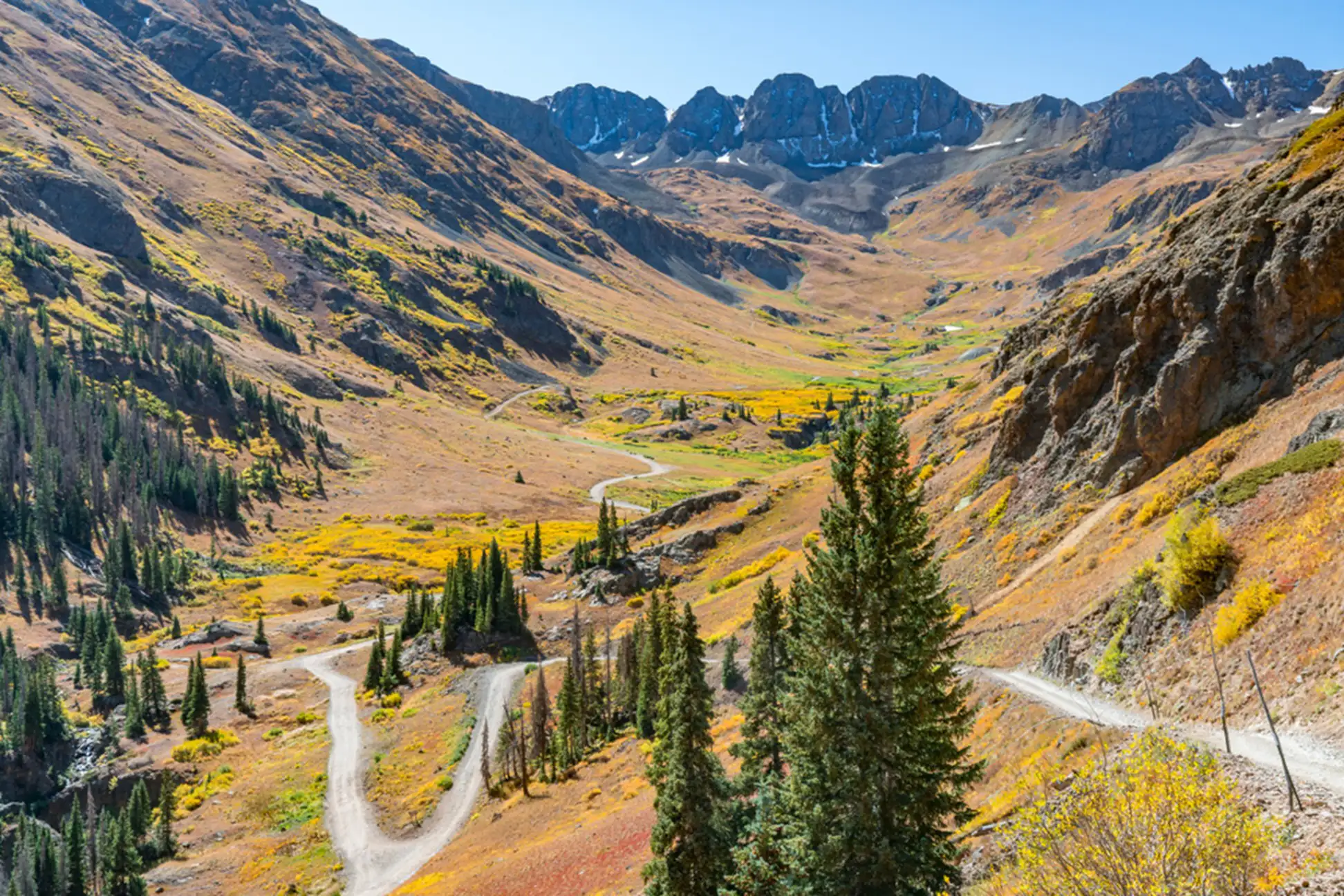 A winding dirt road—one of the epic U.S. trails—cuts through a colorful mountain valley with scattered evergreen trees, yellow autumn foliage, and rugged peaks in the background under a clear blue sky.