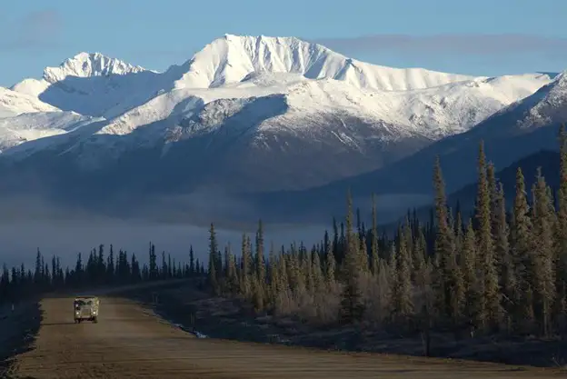 A vehicle with an overland trailer drives on a dirt road lined with tall evergreen trees, with snow-capped mountains rising in the background under a blue U.S. sky—perfect for those seeking epic trails.