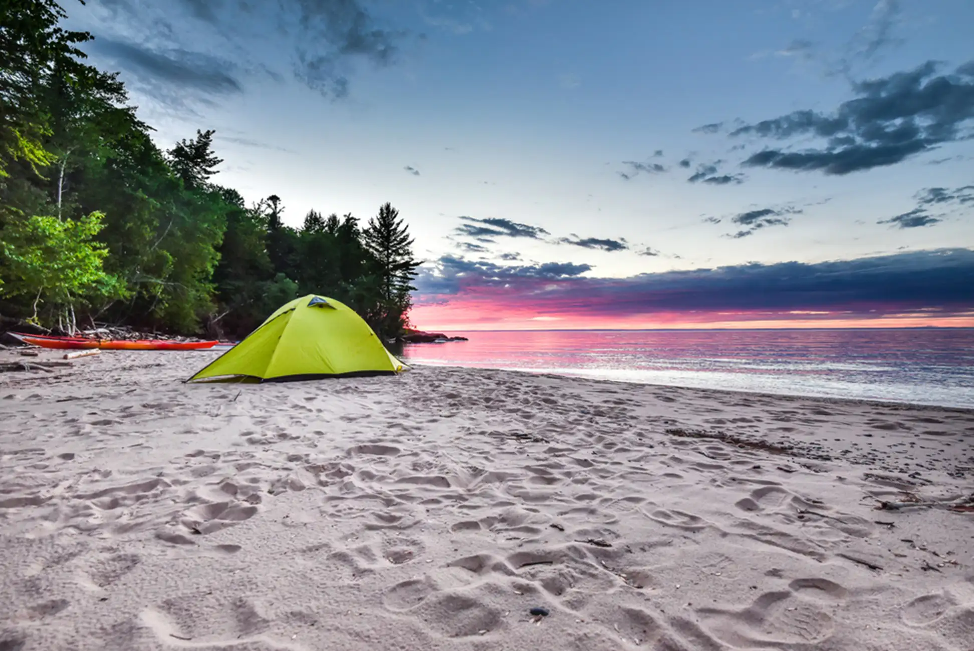 A bright green tent sits on a sandy beach near the water's edge, with trees in the background and a colorful sunset sky reflecting on the calm lake—perfect for exploring epic trails across the U.S.