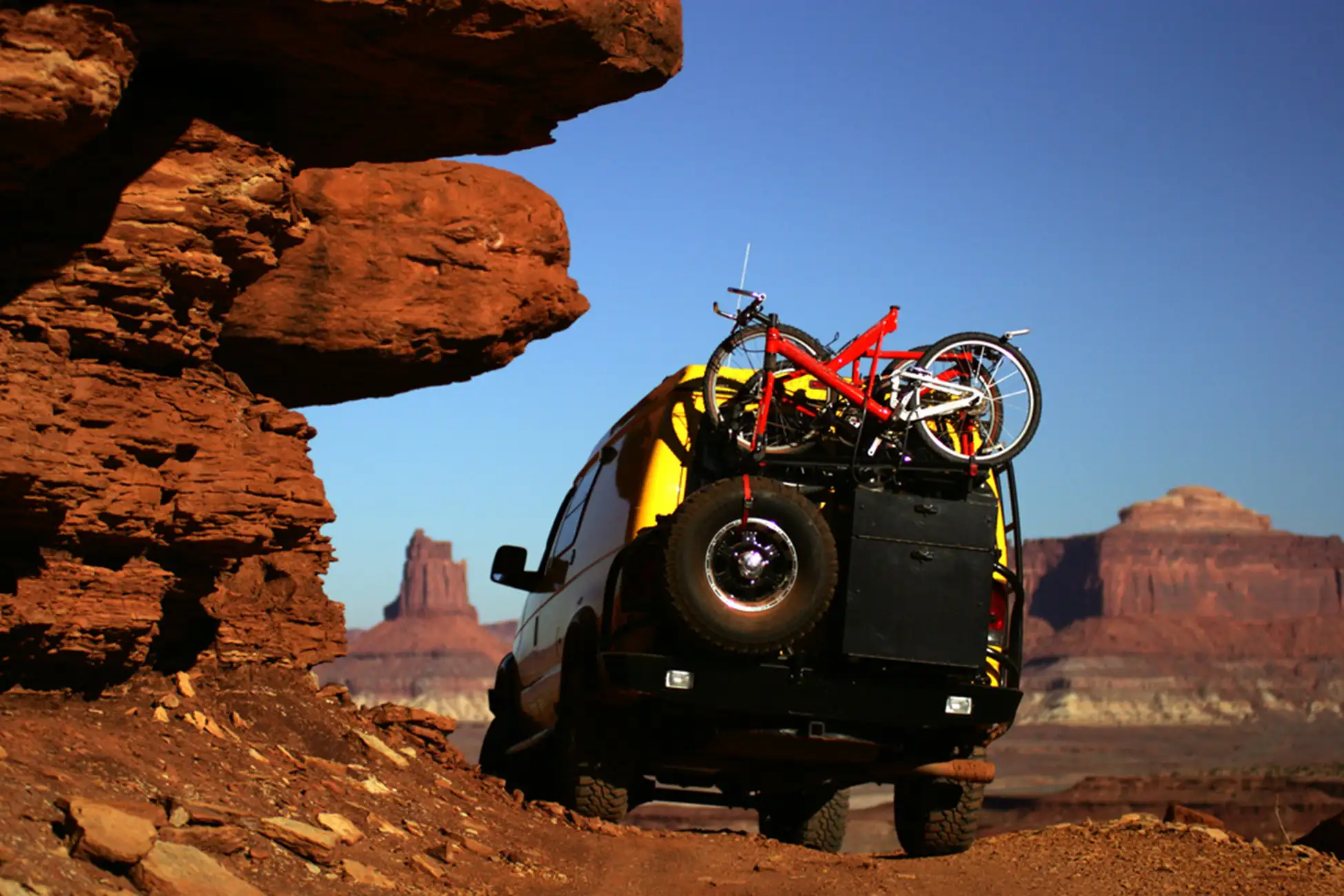 A van with two bicycles mounted on the back and an overland trailer drives on a dirt road through a desert landscape with large red rock formations under a clear blue sky, heading toward epic trails in the U.S.