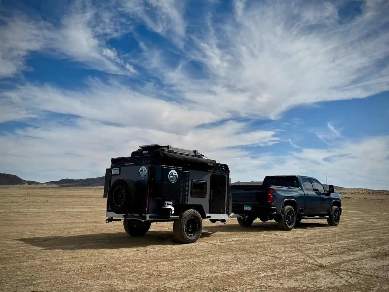 A dark blue pickup truck towing an off-road camper trailer is parked on a dry, cracked desert landscape under a partly cloudy blue sky. Mountains are visible in the distant background.