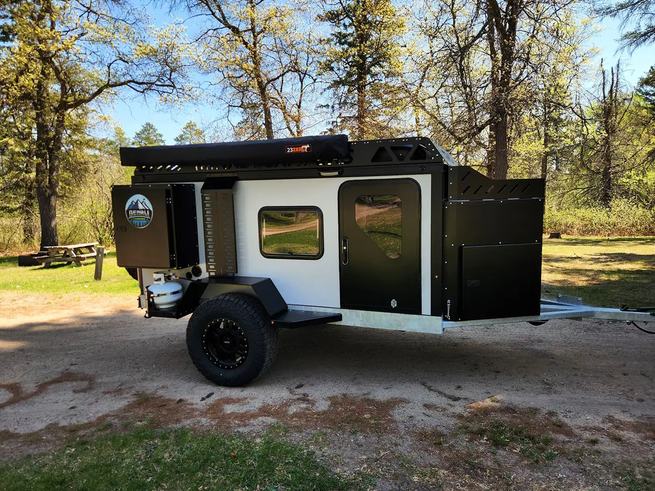 A black and white Square Drop Trailer camper, built for adventures, is parked on a dirt path in a wooded area. Rugged tires and outdoor storage compartments make it perfect for off-road camping among the trees and green grass.