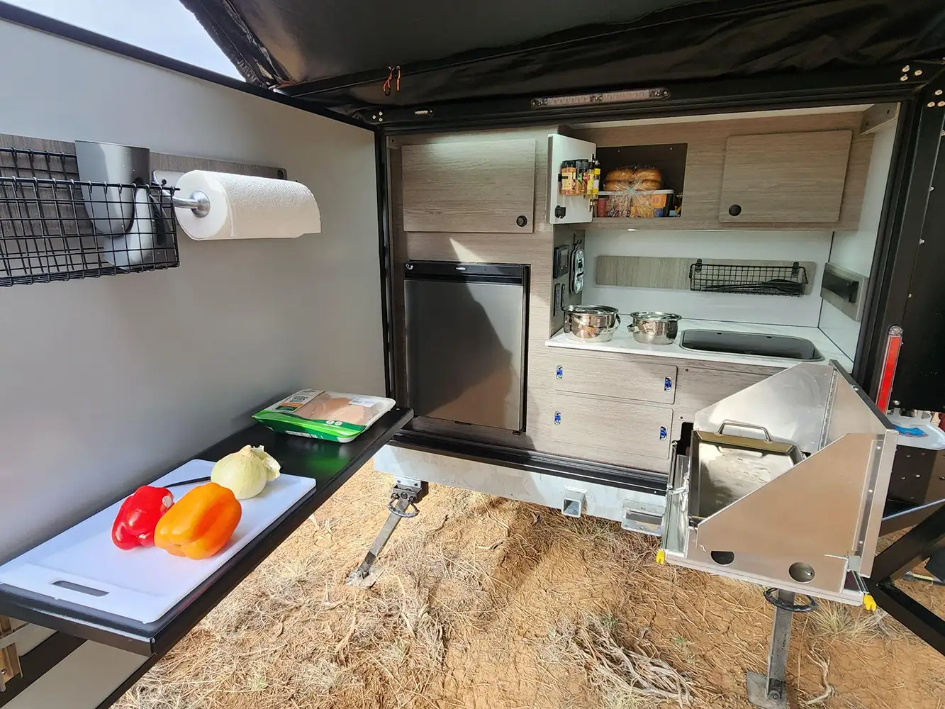 Outdoor camper kitchen with light wood cabinets in a Square Drop Trailer, featuring a small fridge, pots on the stove, a cutting board with chopped peppers and onion, paper towel holder, and kitchen supplies—all set for your next adventures on dry grass.