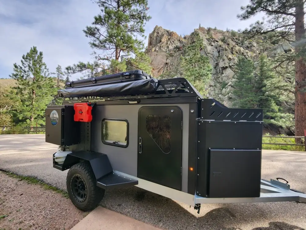 A modern Square Drop Trailer with large tires, roof storage, and outdoor gear is parked on a paved area surrounded by pine trees and rocky cliffs—a perfect camper for outdoor adventures under a clear sky.