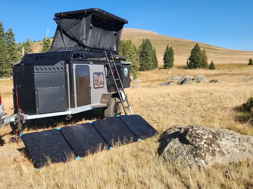 A rugged Square Drop Trailer with an extended pop-up tent is parked on dry grassland, near trees and hills. Solar panels set up in front of the camper harness sunlight for power, ready for adventures in the wild.
