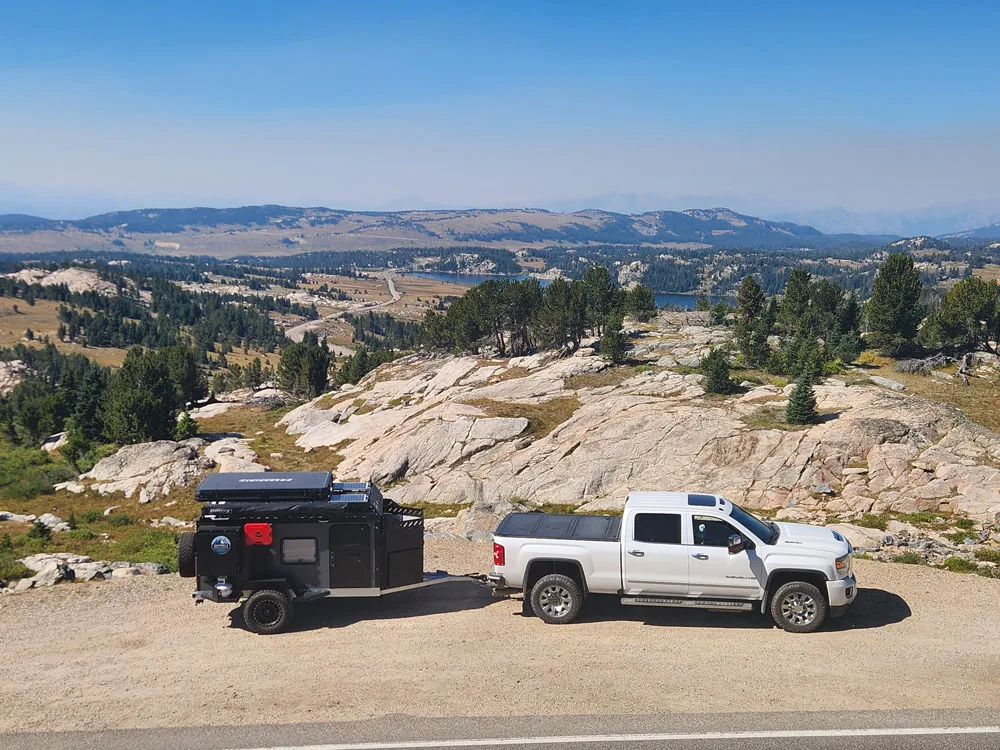 A white pickup truck towing a black Square Drop Trailer is parked on the roadside, overlooking a scenic, rocky landscape with distant hills, trees, and blue sky—perfect for adventures.