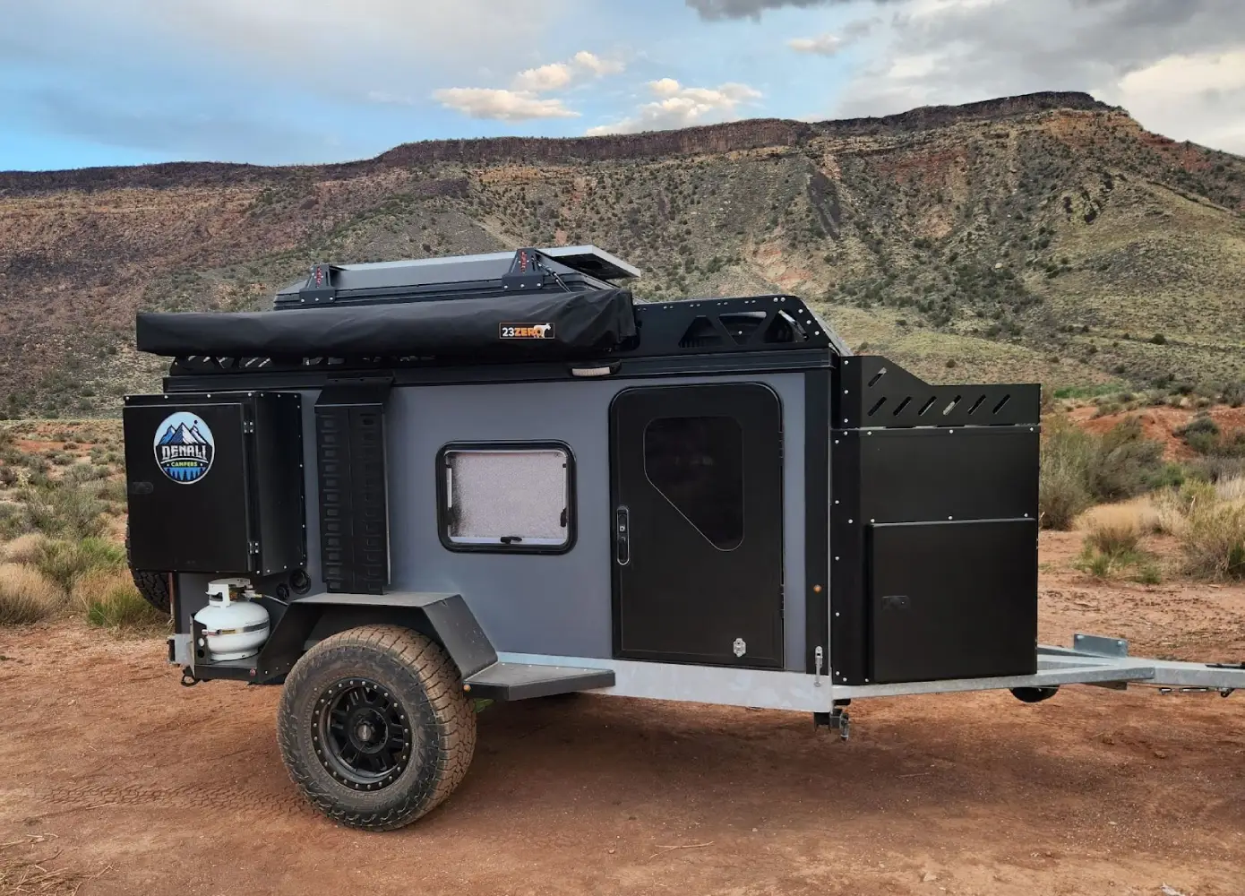 A small, black off-road camping trailer with a roof rack and side awning is parked on rocky desert ground, with red cliffs and sparse vegetation in the background under a partly cloudy sky.