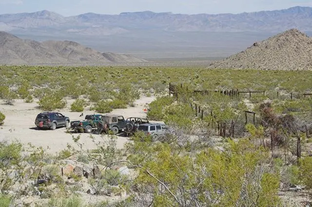Several off-road vehicles are parked on a sandy desert landscape surrounded by sparse bushes and hills, with mountains visible in the distance under a clear sky.