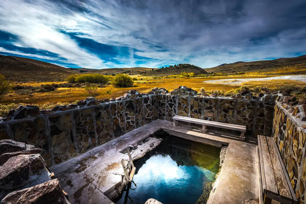 A small stone-walled hot spring pool with clear water sits in a rural landscape of yellow grass and rolling hills under a partly cloudy blue sky. Wooden benches line the edge of the pool.