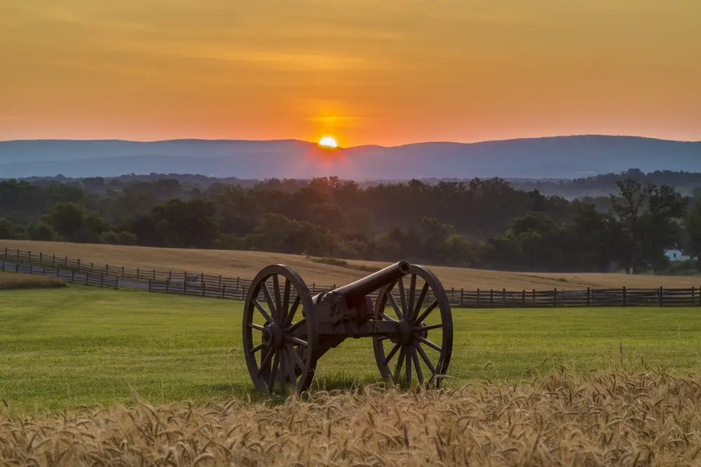 An old cannon stands in a grassy field at sunrise, with rolling hills and a wooden fence in the background, and the sun rising over the distant mountains under an orange sky.